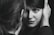 A close-up black and white photograph capturing a woman gazing intently at the camera, framed by a man's hand gently holding her face. The focus is on her expressive eyes and subtle smile, creating an intimate and personal atmosphere.