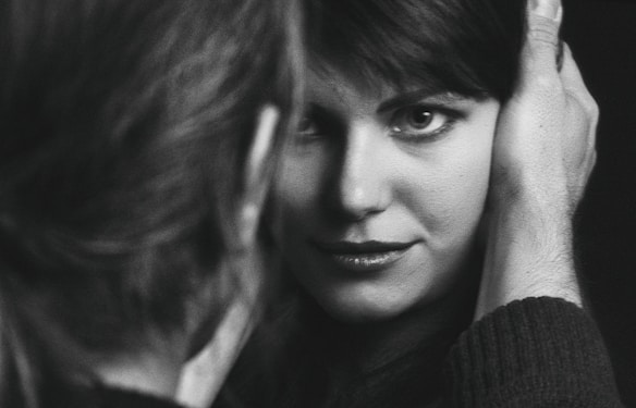 A close-up black and white photograph capturing a woman gazing intently at the camera, framed by a man's hand gently holding her face. The focus is on her expressive eyes and subtle smile, creating an intimate and personal atmosphere.