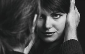 A close-up black and white photograph capturing a woman gazing intently at the camera, framed by a man's hand gently holding her face. The focus is on her expressive eyes and subtle smile, creating an intimate and personal atmosphere.
