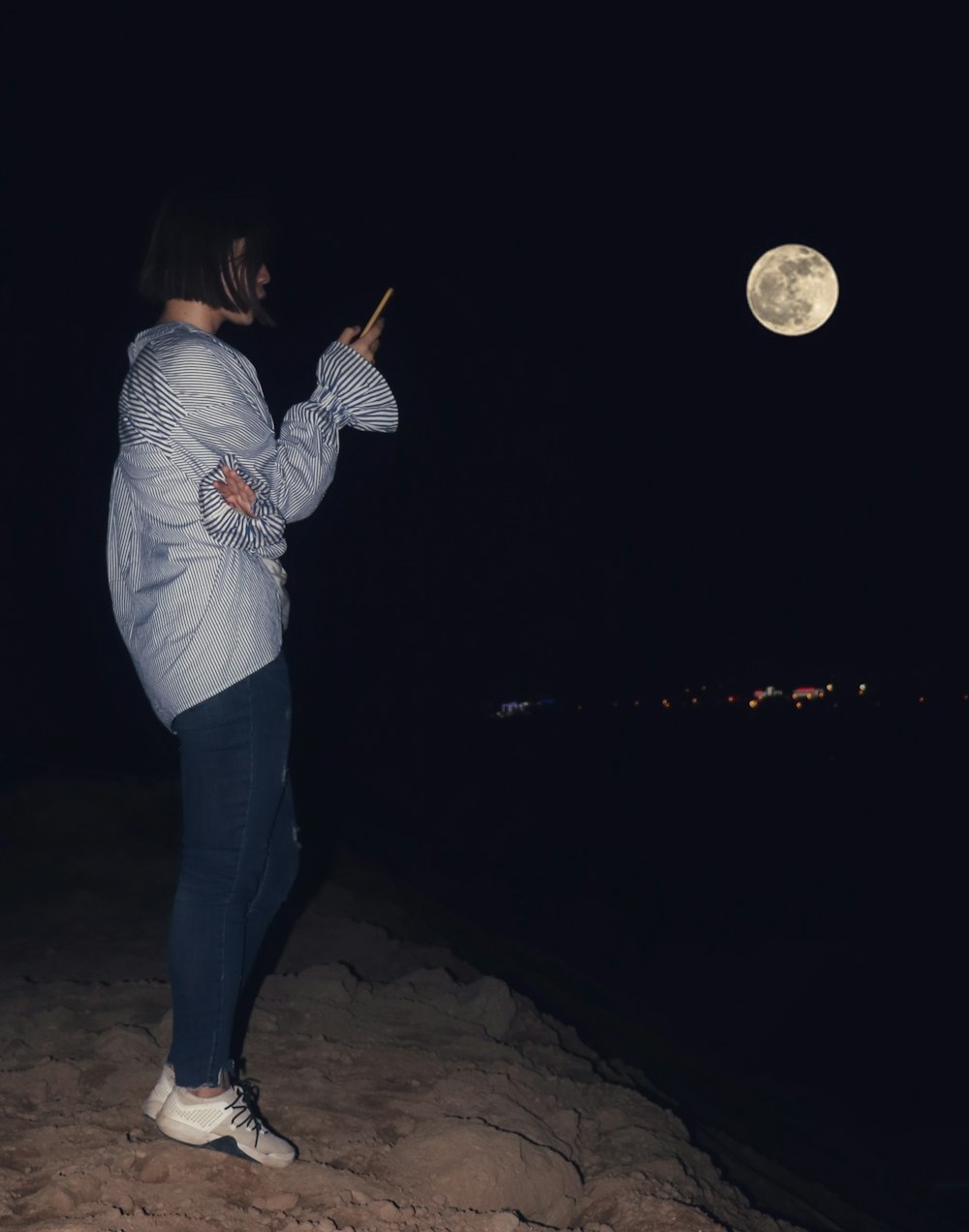 woman wearing purple blouse and black jeans standing on soil surface while using smartphone during nighttime