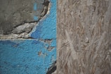 A close-up view of a weathered wall with two distinct textures: on the left, cracked and peeling blue paint on a rough concrete surface, and on the right, a section of oriented strand board with a natural wood texture.
