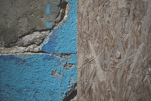 A close-up view of a weathered wall with two distinct textures: on the left, cracked and peeling blue paint on a rough concrete surface, and on the right, a section of oriented strand board with a natural wood texture.