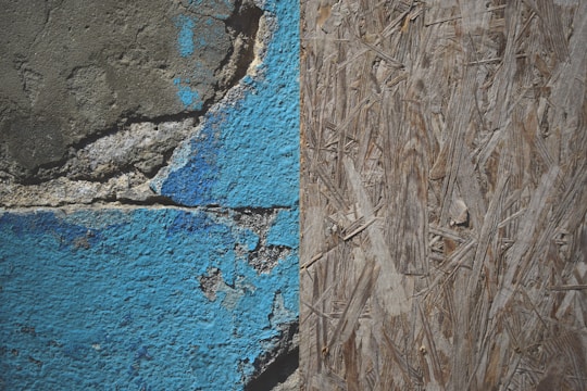 A close-up view of a weathered wall with two distinct textures: on the left, cracked and peeling blue paint on a rough concrete surface, and on the right, a section of oriented strand board with a natural wood texture.