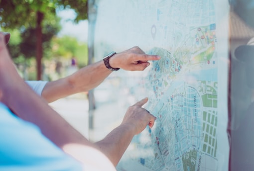 Hands are pointing at a detailed city map with various marked locations and streets. The map is outdoors, with greenery visible in the blurred background.
