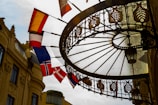 A wrought iron and glass canopy extends over a walkway, with ornamental detailing and a hanging lantern. Several flags, including the flag of Norway, are hanging from poles on the side of a historic building with classical architectural details. The sun creates a backlight effect, casting shadows through the glass.