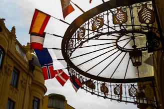 A wrought iron and glass canopy extends over a walkway, with ornamental detailing and a hanging lantern. Several flags, including the flag of Norway, are hanging from poles on the side of a historic building with classical architectural details. The sun creates a backlight effect, casting shadows through the glass.