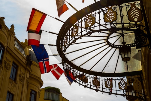 A wrought iron and glass canopy extends over a walkway, with ornamental detailing and a hanging lantern. Several flags, including the flag of Norway, are hanging from poles on the side of a historic building with classical architectural details. The sun creates a backlight effect, casting shadows through the glass.
