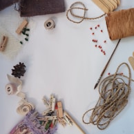 A collection of craft materials including twine, a paintbrush, wooden clothespins, fabric swatches, a pinecone, red beads, and a roll of tape are arranged on a white surface.