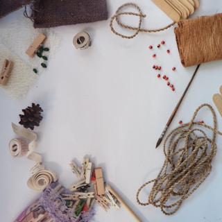 A collection of craft materials including twine, a paintbrush, wooden clothespins, fabric swatches, a pinecone, red beads, and a roll of tape are arranged on a white surface.