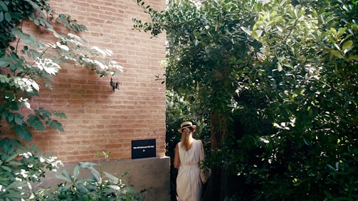 A person wearing a hat and a white dress walks through a lush, green garden beside a brick wall. Sunlight filters through the leaves, casting dappled shadows. A small black sign on the wall reads 'We will show you the way' with an arrow pointing in the direction the person is walking.