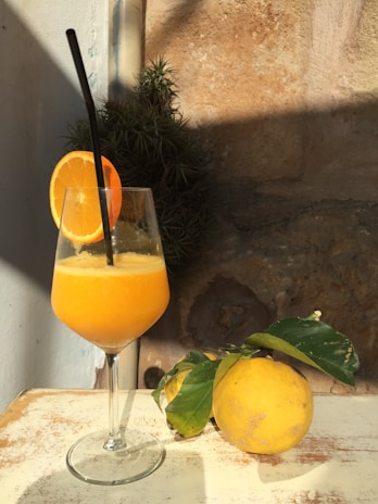 A glass filled with a vibrant orange juice, garnished with a slice of orange and a black straw, is placed on a light-colored surface. Next to the glass, there is a whole citrus fruit with green leaves still attached. The background consists of textured stone walls with sunlight casting shadows.