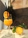 A glass filled with a vibrant orange juice, garnished with a slice of orange and a black straw, is placed on a light-colored surface. Next to the glass, there is a whole citrus fruit with green leaves still attached. The background consists of textured stone walls with sunlight casting shadows.