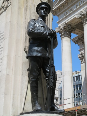 A bronze statue of a soldier in uniform stands proudly, holding a rifle. It is positioned against a grand architectural backdrop with columns and ornate decorations. The sky is clear and blue, adding contrast to the stone and metal elements.