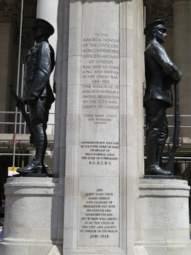 A stone memorial featuring two statues of soldiers standing back to back. Each soldier is dressed in early 20th-century military uniform and holds a rifle. The memorial includes engraved text honoring officers and men from London who served in World War I and mentions the unveiling date, November 12th, 1920. Additional text commemorates those who served in World War II.