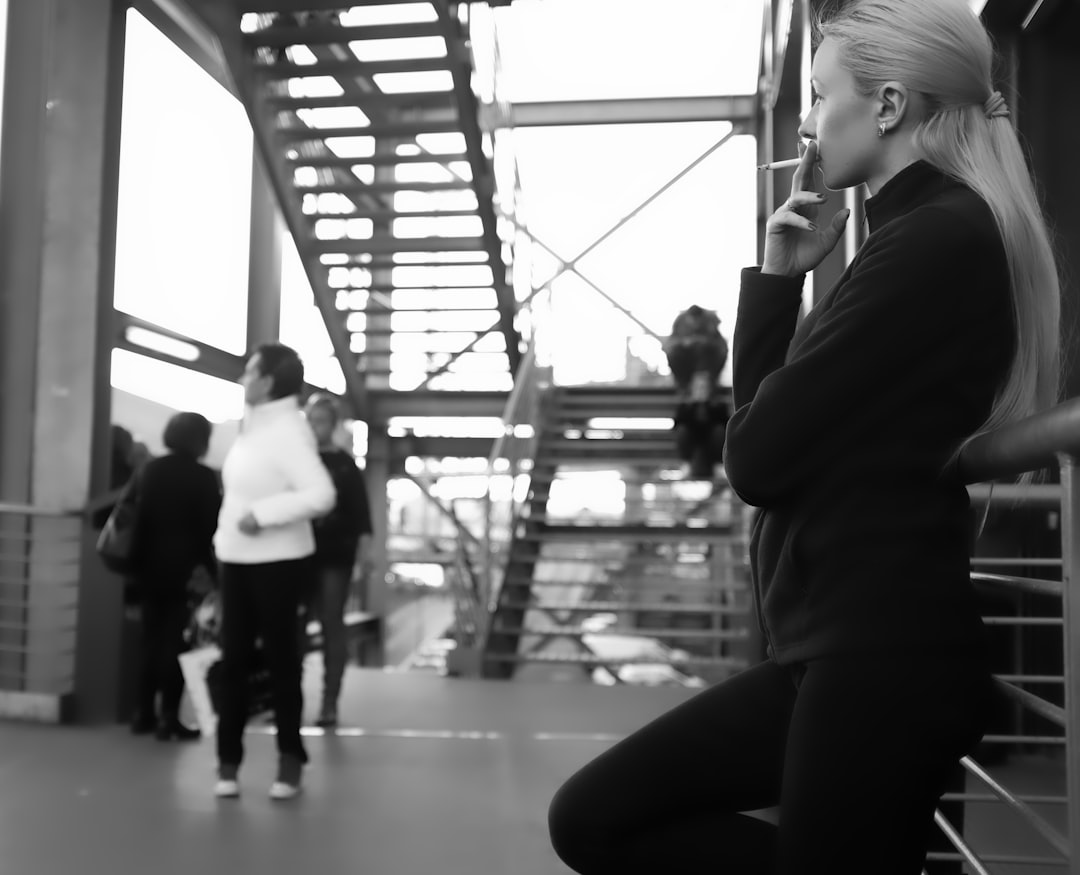 grayscale photography of woman using cigarette while leaning on metal rail