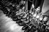 A collection of various pairs of shoes arranged on a wooden floor, featuring a mix of formal and casual styles. The shoes are mostly dark in color, with some lighter pairs visible towards the back. The arrangement suggests a vintage or second-hand shop setting.