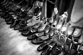 A collection of various pairs of shoes arranged on a wooden floor, featuring a mix of formal and casual styles. The shoes are mostly dark in color, with some lighter pairs visible towards the back. The arrangement suggests a vintage or second-hand shop setting.