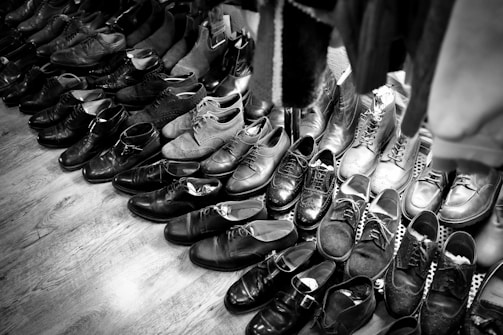 A collection of various pairs of shoes arranged on a wooden floor, featuring a mix of formal and casual styles. The shoes are mostly dark in color, with some lighter pairs visible towards the back. The arrangement suggests a vintage or second-hand shop setting.
