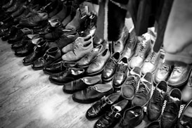 A collection of various pairs of shoes arranged on a wooden floor, featuring a mix of formal and casual styles. The shoes are mostly dark in color, with some lighter pairs visible towards the back. The arrangement suggests a vintage or second-hand shop setting.