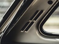 Interior section of a car showing part of the rear window and the rear pillar, including small vents. The materials have a smooth, metallic finish, and natural light is visible coming through the window.