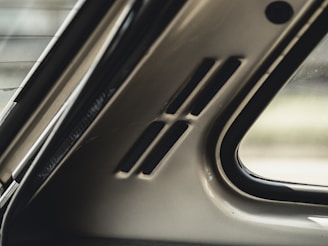 Interior section of a car showing part of the rear window and the rear pillar, including small vents. The materials have a smooth, metallic finish, and natural light is visible coming through the window.