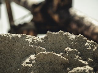 A close-up view of a rough, textured surface resembling concrete or stone. The background features a blurry structure that seems to be part of a machine, likely some form of construction equipment. The light captures the crevices and shadows in the rock, providing a sense of depth.