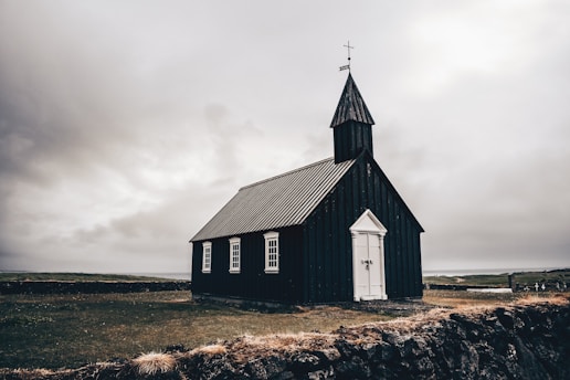 A small, dark-colored church building with a pointed steeple and a cross on top. The church has white-framed windows and a white door, and it is set in a rural, open landscape with a cloudy sky in the background. There is a stone wall in the foreground and some sparse grass surrounding the church.