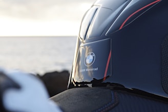 A close-up view of a BMW Motorrad logo on a sleek, dark motorcycle helmet with red accents. The background features an out-of-focus seascape, suggesting a coastal setting during possibly a sunset or sunrise.