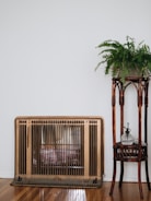 A vintage heater with a bronze finish stands against a white wall, next to a wooden plant stand holding a lush green fern. The wooden floor enhances the cozy, rustic feel.