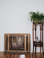 A vintage heater with a bronze finish stands against a white wall, next to a wooden plant stand holding a lush green fern. The wooden floor enhances the cozy, rustic feel.