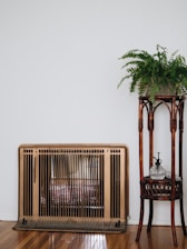 A vintage heater with a bronze finish stands against a white wall, next to a wooden plant stand holding a lush green fern. The wooden floor enhances the cozy, rustic feel.
