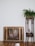 A vintage heater with a bronze finish stands against a white wall, next to a wooden plant stand holding a lush green fern. The wooden floor enhances the cozy, rustic feel.