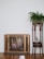 A vintage heater with a bronze finish stands against a white wall, next to a wooden plant stand holding a lush green fern. The wooden floor enhances the cozy, rustic feel.