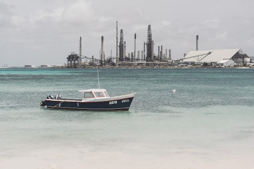 A small boat with the registration A976 is anchored in the calm, turquoise sea near a shoreline. In the background, an industrial facility with multiple chimneys and large structures extends across the horizon, suggesting a refinery or factory setting. A bird is flying above the water, adding to the serene yet industrialized seaside atmosphere.