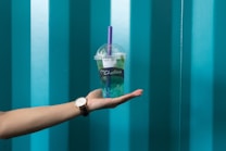 A hand with a watch on the wrist is holding a bubble tea cup with a purple straw. The cup has a brand logo and is partially filled with green liquid. The background features a pattern of vertical teal stripes.