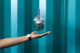 A hand with a watch on the wrist is holding a bubble tea cup with a purple straw. The cup has a brand logo and is partially filled with green liquid. The background features a pattern of vertical teal stripes.