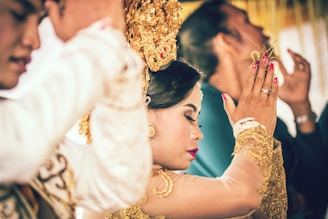 A woman is dressed in traditional attire, adorned with gold accessories and intricate headwear, surrounded by others in a ceremonial setting. The focus is on her serene expression and gesture, suggesting a moment of cultural significance.