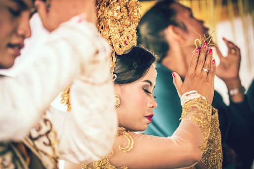 A woman is dressed in traditional attire, adorned with gold accessories and intricate headwear, surrounded by others in a ceremonial setting. The focus is on her serene expression and gesture, suggesting a moment of cultural significance.