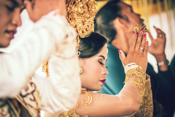A woman is dressed in traditional attire, adorned with gold accessories and intricate headwear, surrounded by others in a ceremonial setting. The focus is on her serene expression and gesture, suggesting a moment of cultural significance.