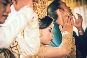 A woman is dressed in traditional attire, adorned with gold accessories and intricate headwear, surrounded by others in a ceremonial setting. The focus is on her serene expression and gesture, suggesting a moment of cultural significance.