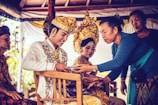 A ceremonial scene featuring individuals in traditional attire. A man wearing an ornate embroidered jacket and a richly decorated headdress smiles while receiving an object from a woman in a blue top. A woman beside him, also wearing an elaborate headdress, looks on, portraying a sense of cultural heritage.