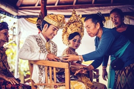 A ceremonial scene featuring individuals in traditional attire. A man wearing an ornate embroidered jacket and a richly decorated headdress smiles while receiving an object from a woman in a blue top. A woman beside him, also wearing an elaborate headdress, looks on, portraying a sense of cultural heritage.