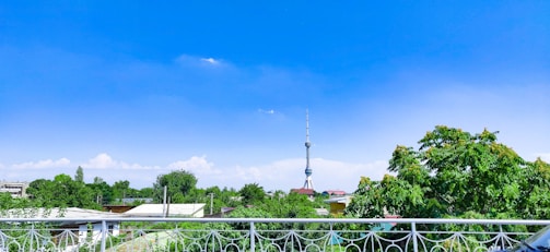 A scenic view featuring a tall broadcasting tower rising into a vibrant blue sky with scattered clouds, surrounded by lush green trees and rooftops.