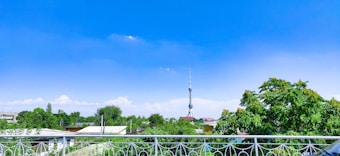 A scenic view featuring a tall broadcasting tower rising into a vibrant blue sky with scattered clouds, surrounded by lush green trees and rooftops.