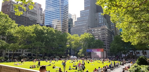 A vibrant city park scene with numerous people relaxing on a green lawn surrounded by tall, modern skyscrapers and lush trees. The sun is shining brightly, casting shadows and illuminating the grass. A large screen is visible in the background, suggesting the organization of some outdoor event or movie.