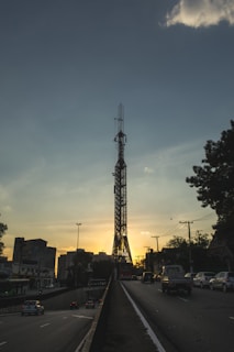 A large metal tower stands prominently against a sunset sky, surrounded by urban infrastructure. Vehicles are driving on a road beneath the tower, with buildings visible on both sides. The scene is framed by silhouetted trees and telecommunication poles.