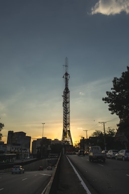 A large metal tower stands prominently against a sunset sky, surrounded by urban infrastructure. Vehicles are driving on a road beneath the tower, with buildings visible on both sides. The scene is framed by silhouetted trees and telecommunication poles.