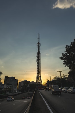 A large metal tower stands prominently against a sunset sky, surrounded by urban infrastructure. Vehicles are driving on a road beneath the tower, with buildings visible on both sides. The scene is framed by silhouetted trees and telecommunication poles.