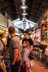 Inside a bustling, covered market with people walking and standing around, observing various stalls filled with goods. The arched ceiling features bright hanging lights, and the scene is lively and energetic with several individuals present.