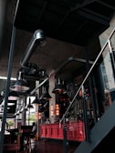 A modern, industrial-style interior with exposed ductwork and large, metallic pendant lights hanging from the ceiling. There is a red-painted bar or counter area, with vertical neon lights illuminating the space. A few tables and chairs are visible, and the area is decorated with various artworks on the wall.
